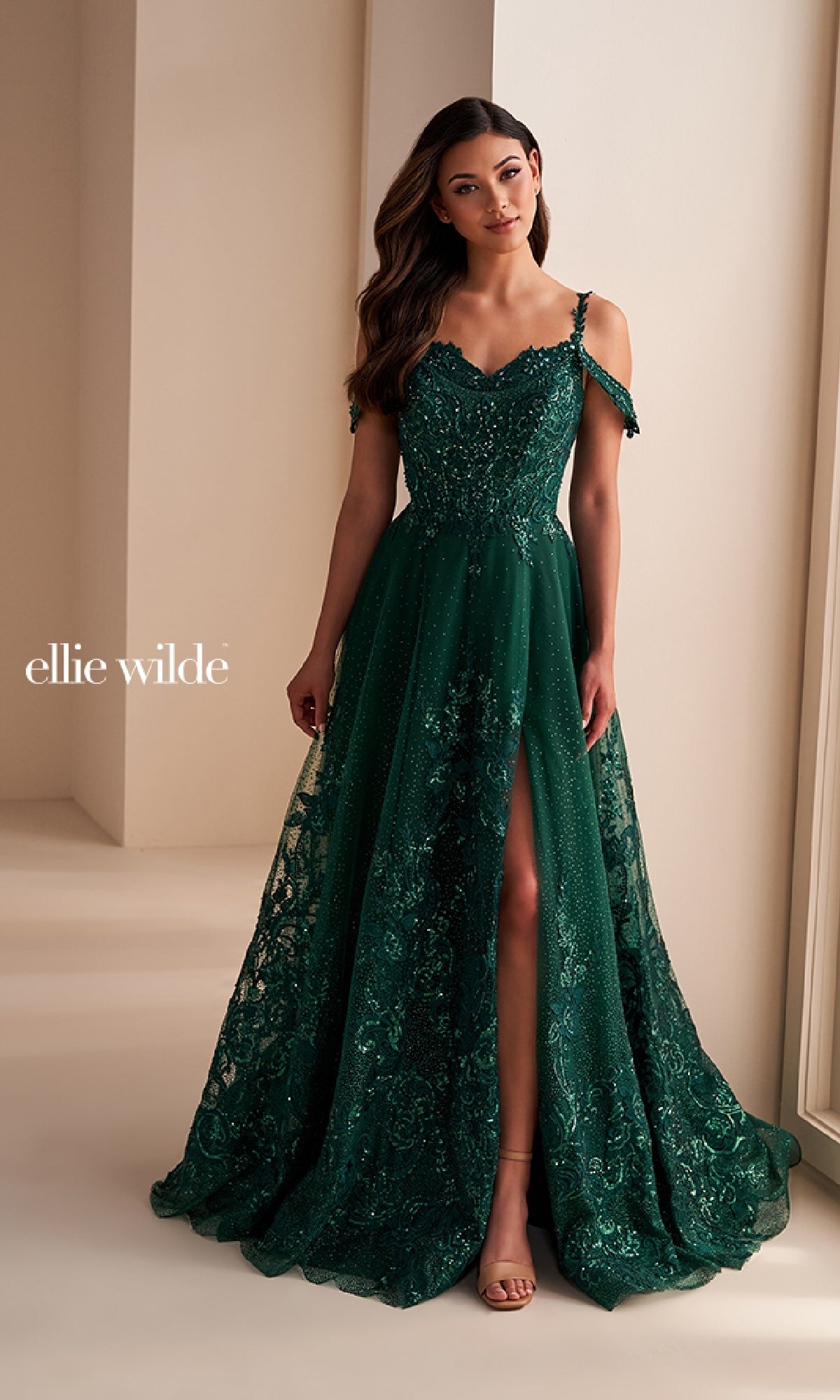 A woman stands indoors wearing the Ellie Wilde EW36220 long prom dress, a dark green off-the-shoulder gown with beaded lace, a front slit, and glitter tulle, paired with her long wavy hair.